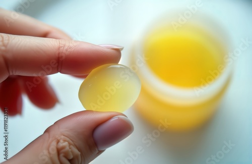 Woman holds light yellow skincare product likely petroleum jelly. Closeup view shows hand applying holding jelly. Product in jar container in background. White background implies studio setting. photo