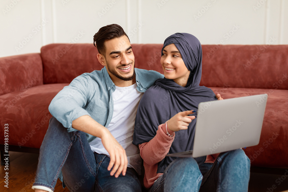 House Ownership And Relocation Project Concept. Portrait Of Young Muslim Family Using Laptop Sitting On Floor After Moving New Home. Spouses Browsing Internet, Woman In Hijab Pointing At Screen House Ownership And Relocation Project Concept. Portrait Of Young Muslim Family Using Laptop Sitting On Floor After Moving New Home. Spouses Browsing Internet, Woman In Hijab Pointing At Screen