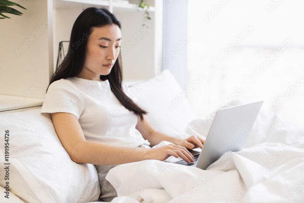 Young Asian woman using laptop on bed in sunlight-filled room, exuding peaceful and focused atmosphere, highlighting casual comfort and technology usage in everyday life. Young Asian woman using laptop on bed in sunlight-filled room, exuding peaceful and focused atmosphere, highlighting casual comfort and technology usage in everyday life.