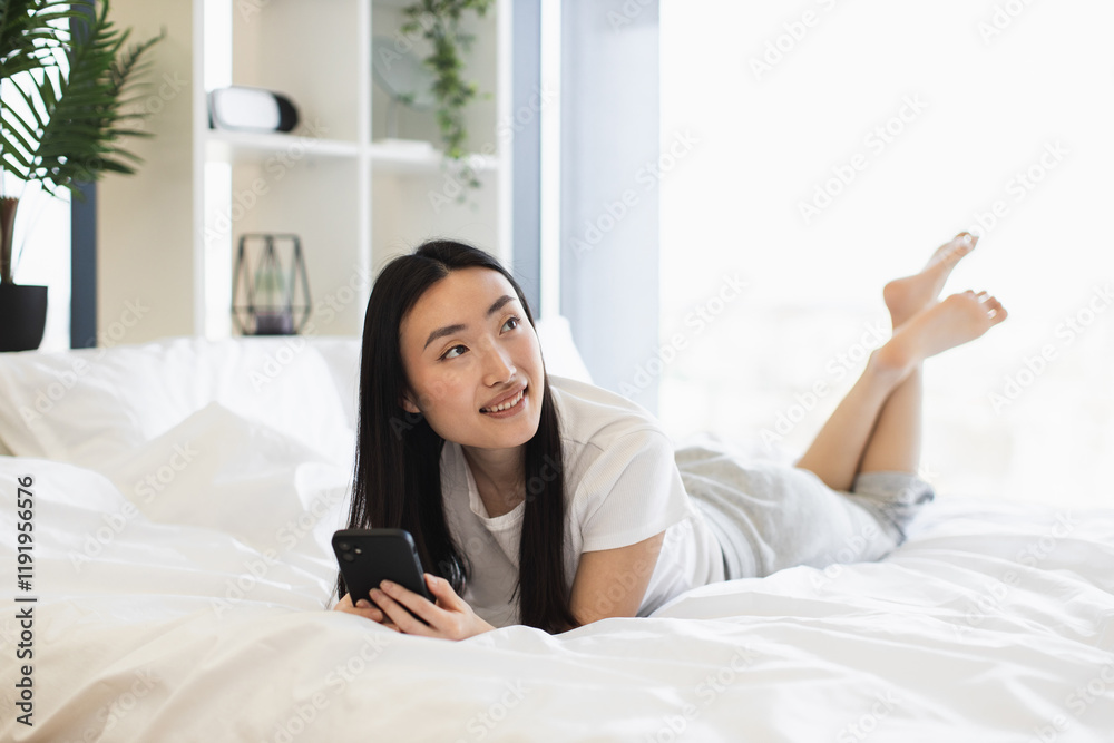 Young Asian woman using smartphone while lying on bed in cozy bright bedroom. Enjoying leisure time, communicating online. Relaxing and comfortable home environment with natural light.
