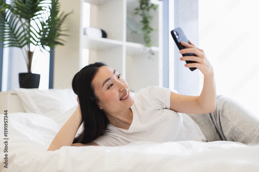 Asian woman in twenties having online video chat on bed, smiling and relaxed in bright room. Asian woman in twenties having online video chat on bed, smiling and relaxed in bright room.