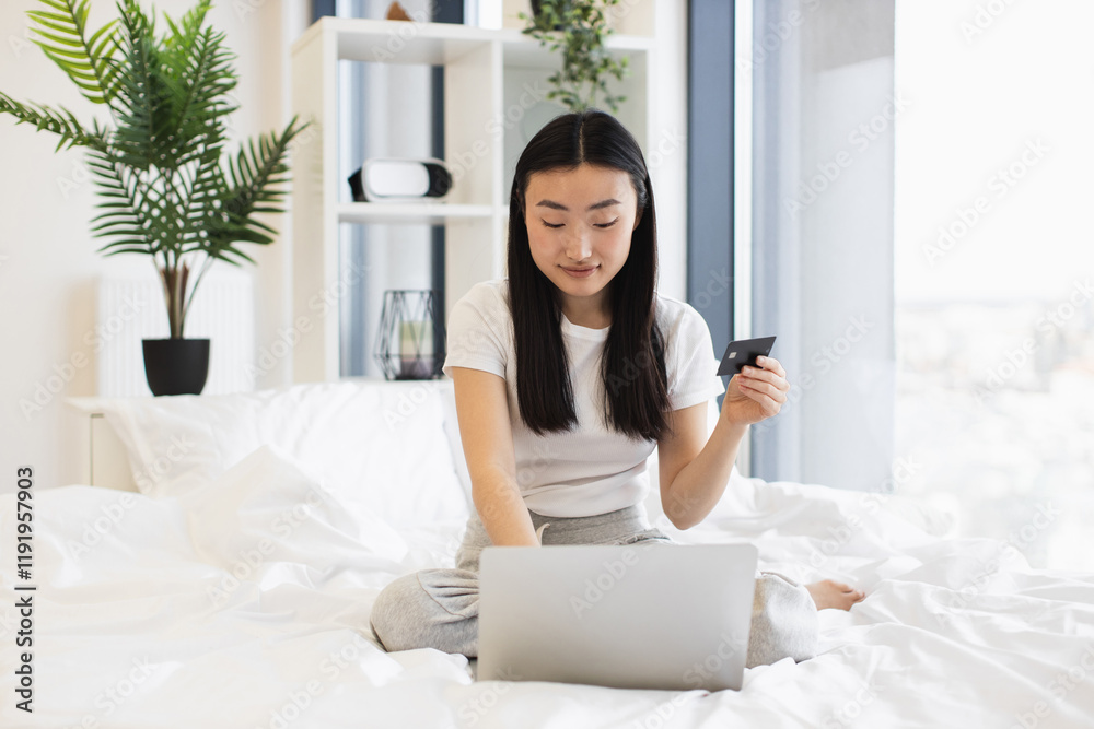 Young Asian woman uses credit card for online shopping on laptop while sitting on bed. Modern technology, convenience, and internet shopping concept.