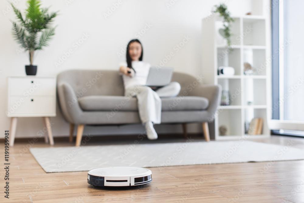 Asian woman relaxing on sofa uses robot vacuum cleaner for home cleaning. Image shows modern living room setup with automated technology. Young adult woman enjoys convenience. Asian woman relaxing on sofa uses robot vacuum cleaner for home cleaning. Image shows modern living room setup with automated technology. Young adult woman enjoys convenience.