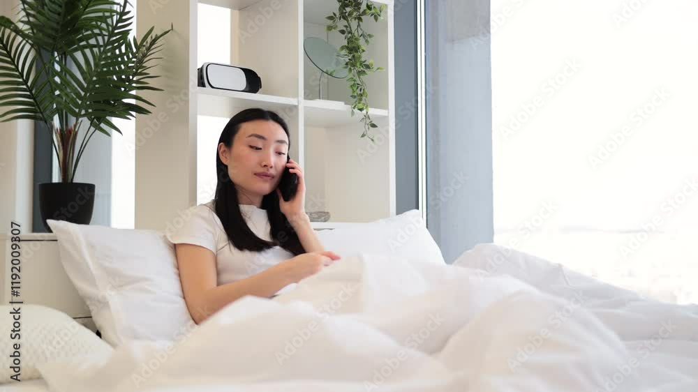 Asian woman in casual white shirt having phone call while relaxing on comfortable bed in bright room. Asian woman in casual white shirt having phone call while relaxing on comfortable bed in bright room.