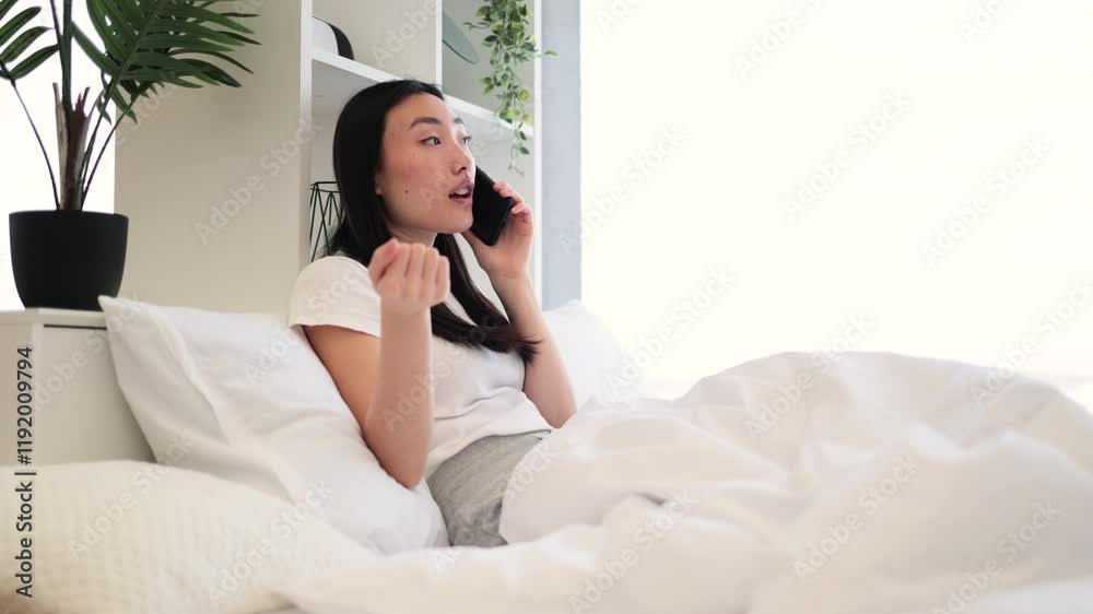 Asian woman in casual white shirt having phone call while relaxing on comfortable bed in bright room. Asian woman in casual white shirt having phone call while relaxing on comfortable bed in bright room.
