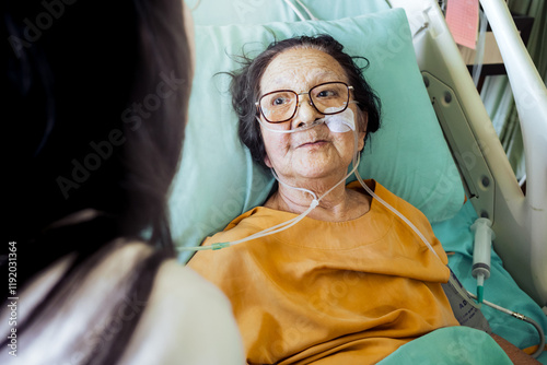 Sick Southeast Asian old female patient in hospital bed with oxygen support wearing eyeglasses seems happy and passionate of being visited by family