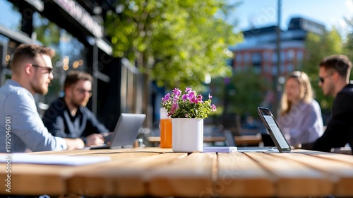 Four young professionals collaborate on laptops at an outdoor cafe table.  Flowers and drinks are on the table.  Sunny day.