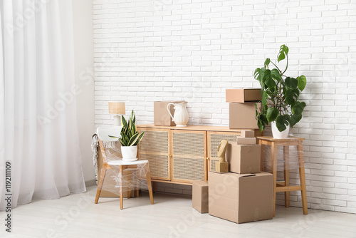 Wooden cabinet  packed chair  houseplants and cardboard boxes near white brick wall. Moving house concept