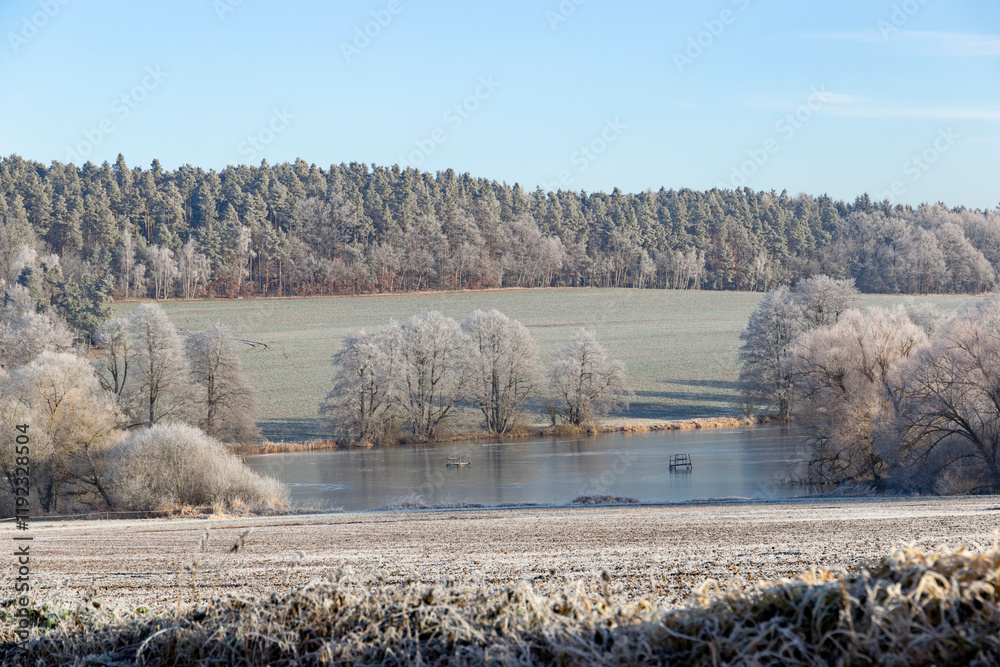 A serene winter morning in the countryside with frost-covered trees, a calm pond, and distant farmhouses under a clear blue sky.