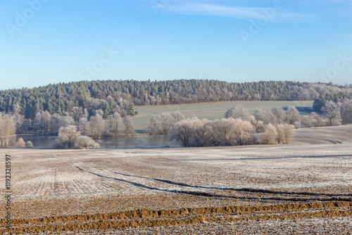 Obraz na płótnie A frosty winter morning in the countryside, featuring a plowed field with visible tire tracks in the foreground, a frozen pond surrounded by bare trees in the middle. w salonie