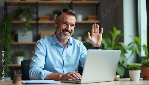 A middle-aged Caucasian man with short dark hair wearing a light blue shirt, waving and smiling while working on a laptop in an office setting