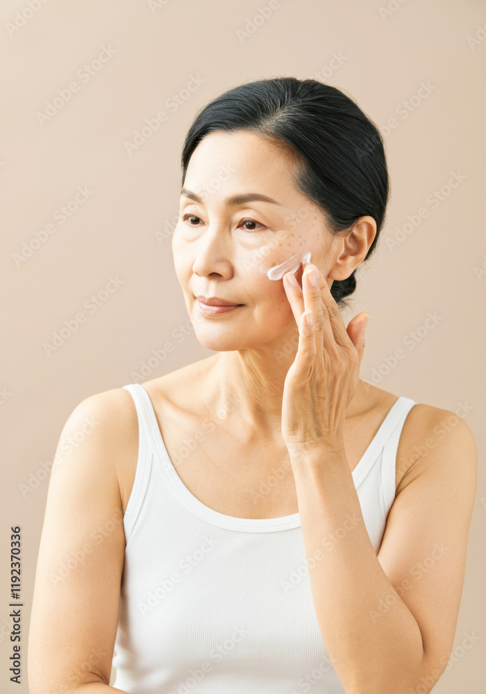 Asian mature female applying skincare cream on face with focused expression Asian mature female applying skincare cream on face with focused expression