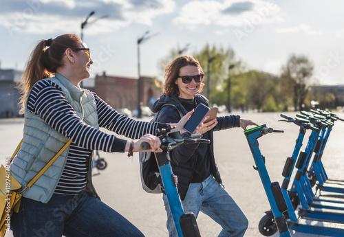Obraz na płótnie Two young women friends using an app to rent a scooter in the city. Active lifestyle w salonie
