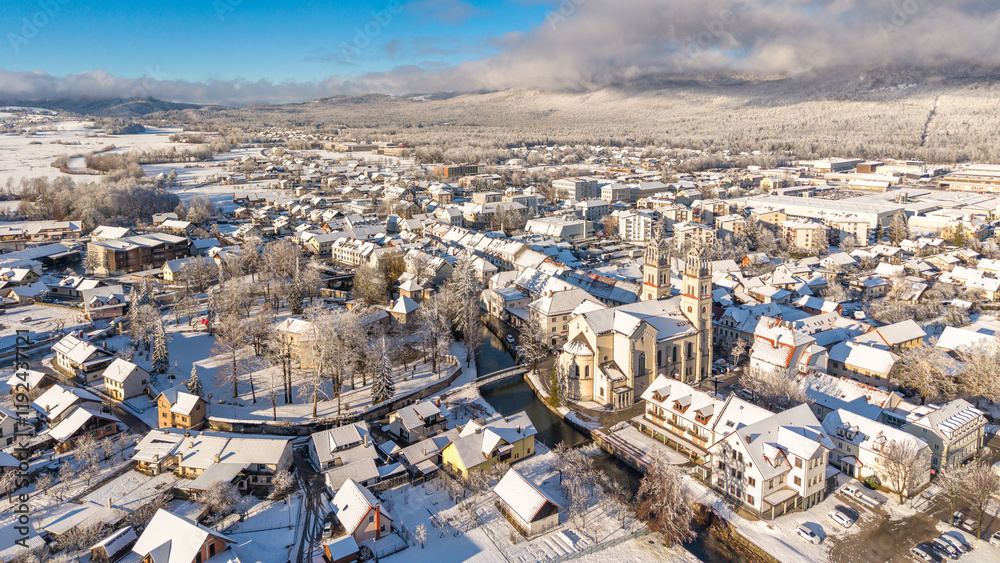 Obraz premium Aerial winter view of Ribnica, Slovenia, with snowy rooftops, St. Stephen's Church, Bistrica River, and a picturesque landscape. Perfect for showcasing European winter charm and tourism
