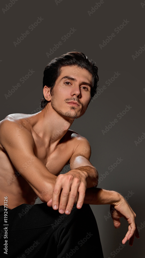 Fototapeta premium Man leaning forward with arms resting on his knees, looking intently into lens. Clean backdrop emphasizes his well-defined features and poised demeanor. Concept of male beauty and facial care,