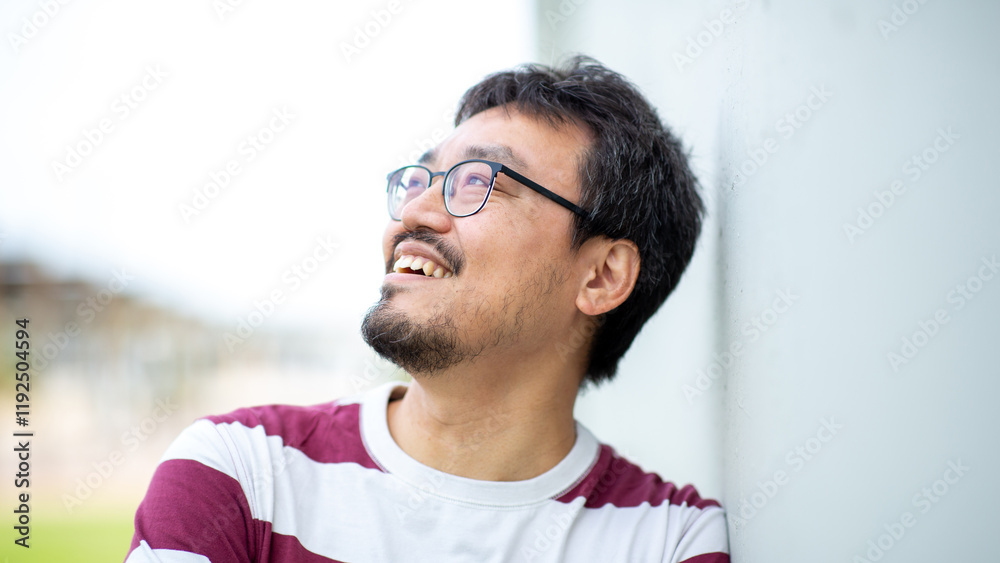 Portrait of an Asian man with glasses smiling and looking up against a white wall Portrait of an Asian man with glasses smiling and looking up against a white wall