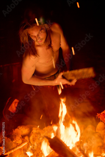 Man with long hair tending to a fire at night, glowing embers illuminating his focused expression. photo