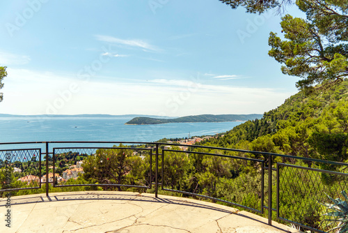 Scenic overlook with a metal railing on a sunny day offering a stunning view of a calm blue adriatic sea, green trees, and a distant coastline, Split, Croatia
