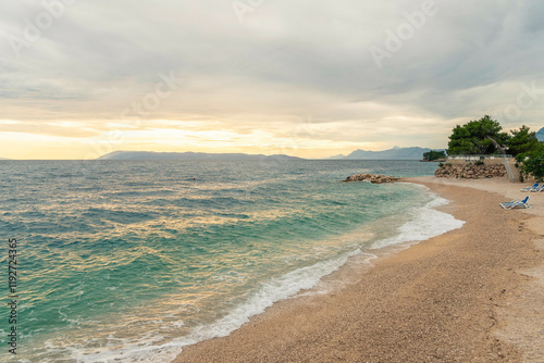 A tranquil beach scene by the Adriatic Sea under a cloudy sky with gentle waves washing ashore and a tree line on the right, Podgora, Croatia