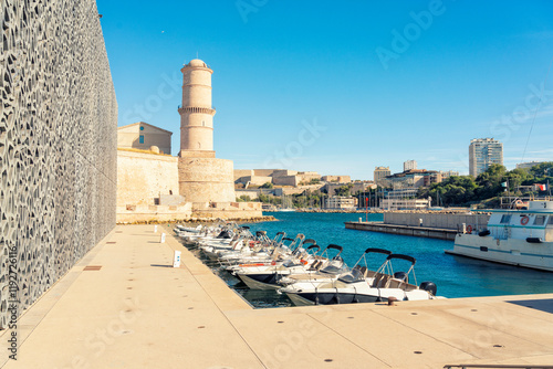 Sunny day view of a historic port of Marseille marina with boats docked beside a modern building adjacent to an old fort of Saint Jean with a watchtower, Marseille, France
