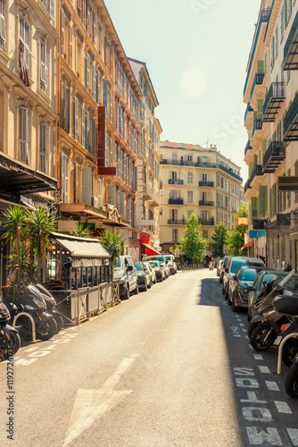 Sunny city street with outdoor cafe seating, lined with traditional European architecture and parked vehicles under a clear blue sky in Nice, France photo