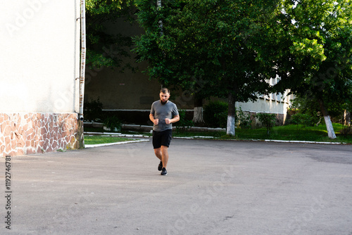 A bearded man in dark shorts and a gray t-shirt jogs along a paved path near a building with trees in the background. He appears focused on his exercise. photo