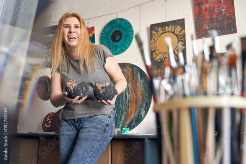 Caucasian female artist showcasing art in studio with paintings and brushes displayed in creative environment.