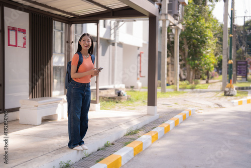 Happy asian university student is standing at bus stop, holding smartphone and waiting for bus, wearing orange t-shirt, blue jeans and backpack