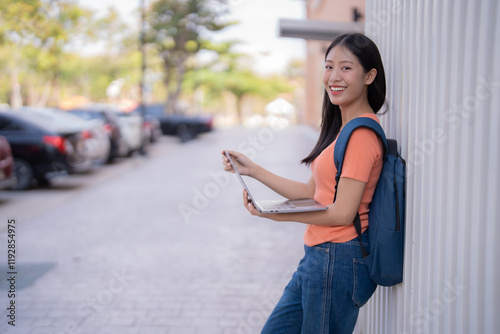 Young Asian student holding a laptop and smiling brightly in the university parking lot after class on a sunny day, enjoying the vibrant campus atmosphere and embracing her academic journey