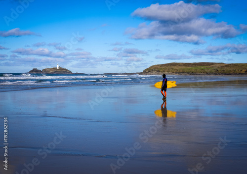 Surfs up on St Ives bay a surfer makes his way to the ocean in a wetsuit and a board.  A reflection is in the wet sand along with  the clouds. Godrevy Lighthouse is seen in the background