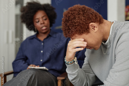 Side view closeup of Black teenage boy crying during support group meeting with woman therapist comforting him in blurred background copy space