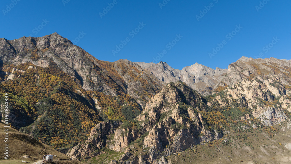 Chegem Gorge. Kabardino-Balkaria. El-Tyubu. Unique rocky ridge with steep cliffs against blue autumn sky.Rocky mountains with trees growing on their slopes glow in morning sun. Chegem Gorge. Kabardino-Balkaria. El-Tyubu. Unique rocky ridge with steep cliffs against blue autumn sky.Rocky mountains with trees growing on their slopes glow in morning sun.