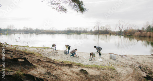 Camera view from far away of group of people gathering trash from beach of big lake or river. Walking around while picking up plastic waste left by others. Volunteering in free time. photo