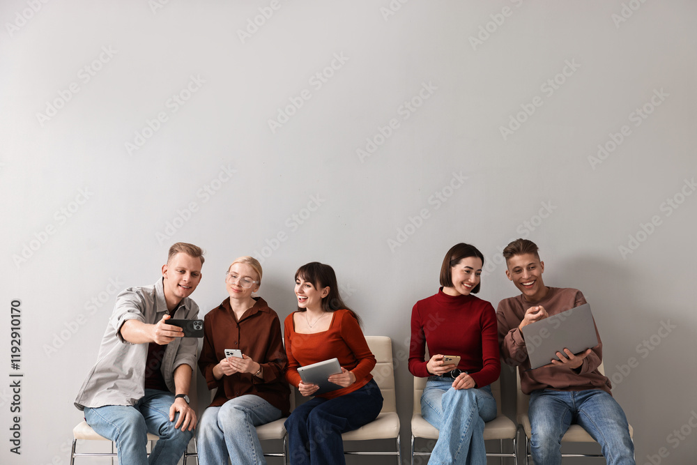 Fototapeta premium Group of people using different gadgets on chairs near light grey wall indoors. Modern technology