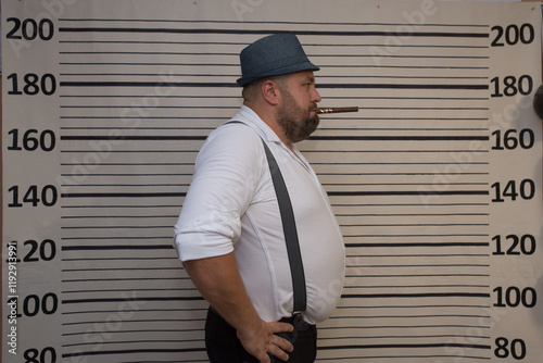 Gangster Party, A man dressed in classic gangster attire, featuring a white shirt, suspenders, and a fedora, poses sternly in front of a height chart with a cigar , completing the mobster look photo