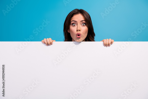 Surprised young woman peeking over white blank banner against blue background, with expressive astonishment and vibrant colors photo