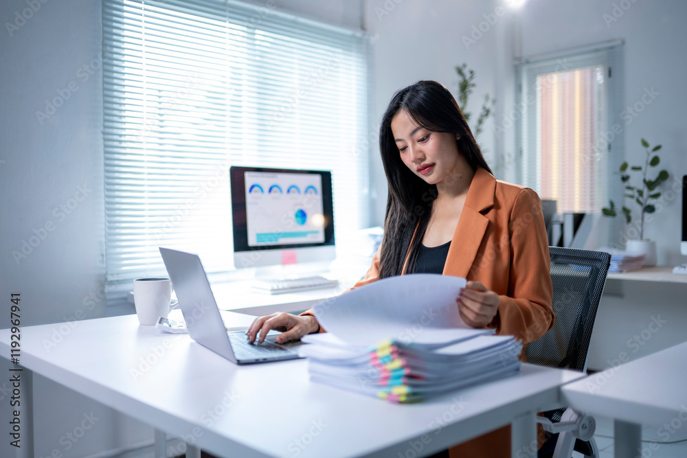 A woman is sitting at a desk with a laptop and a stack of papers