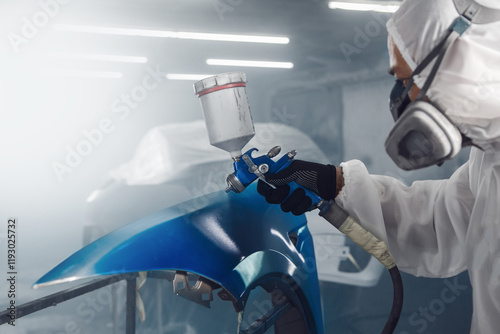 Male Industrial worker sprays paint using airbrush gun on part of car wears in protective mask and overall clothes in workshop garage. photo