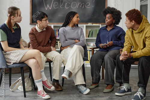 Full length front view portrait of multiethnic group of children with female therapist sitting on chairs in circle during group therapy session in school photo