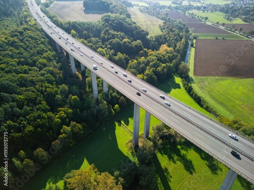 aerial view of a road bridge in Germany, running through a picturesque valley. The bridge is supported by tall concrete pillars that span green fields and forest. Rural settlements and small towns are