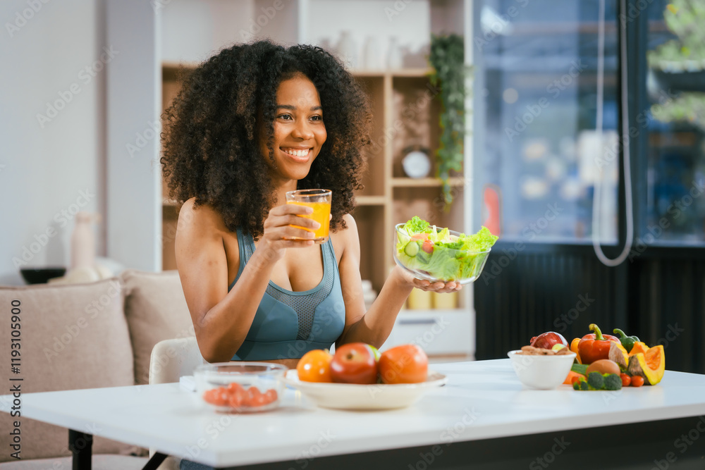 An African American woman in sportswear sits on a sofa near the dining table, preparing a healthy salad with vegetables fruits, drinking juice after exercise for weight control and low-fat nutrition An African American woman in sportswear sits on a sofa near the dining table, preparing a healthy salad with vegetables fruits, drinking juice after exercise for weight control and low-fat nutrition