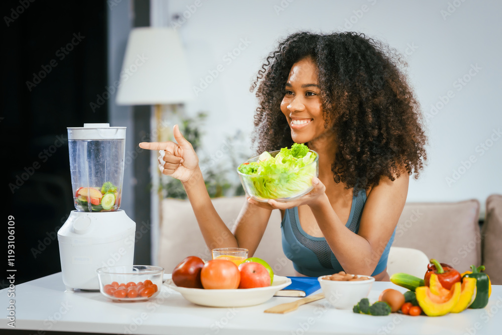 An African American woman in sportswear sits on a sofa near the dining table, preparing a healthy salad with vegetables fruits, drinking juice after exercise for weight control and low-fat nutrition An African American woman in sportswear sits on a sofa near the dining table, preparing a healthy salad with vegetables fruits, drinking juice after exercise for weight control and low-fat nutrition