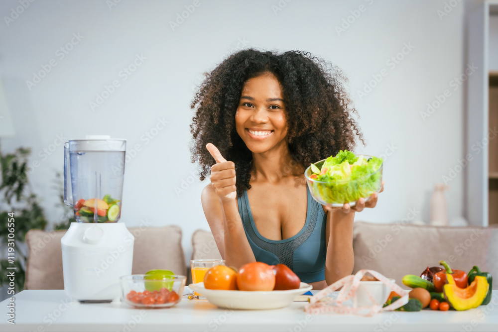 An African American woman in sportswear sits on a sofa near the dining table, preparing a healthy salad with vegetables fruits, drinking juice after exercise for weight control and low-fat nutrition An African American woman in sportswear sits on a sofa near the dining table, preparing a healthy salad with vegetables fruits, drinking juice after exercise for weight control and low-fat nutrition