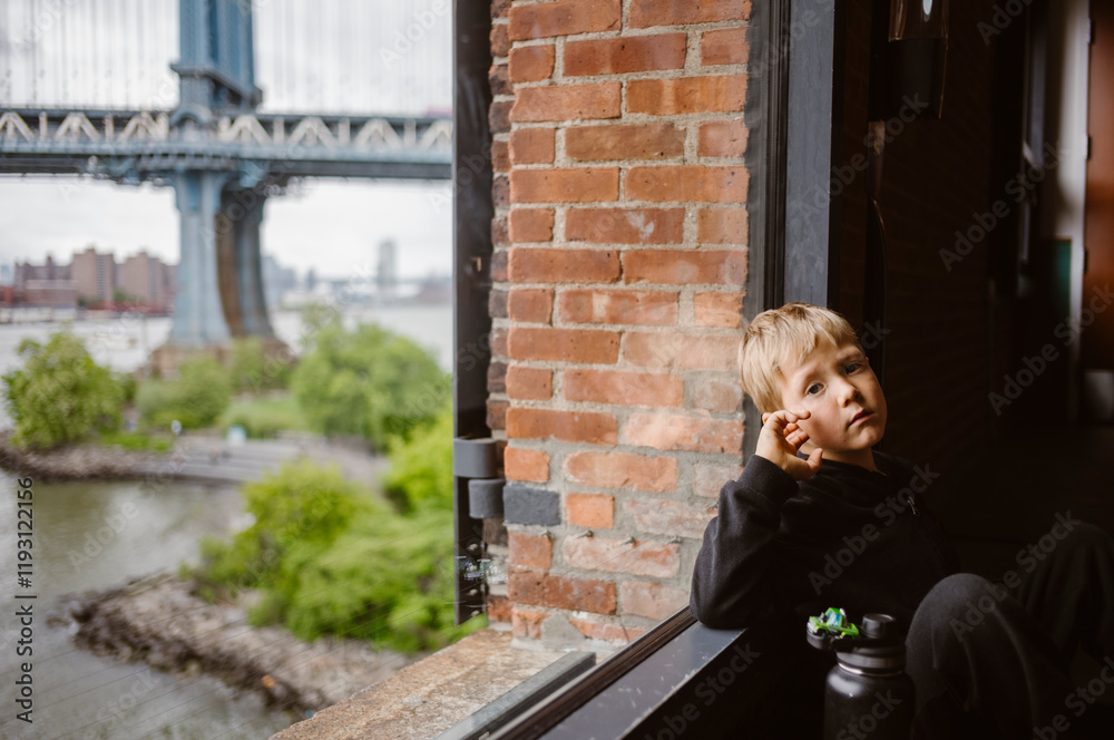 Sad Boy close to window with Manhattan Bridge in the background, in New York City - USA