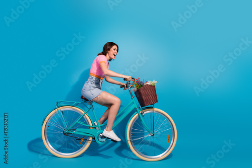 Young woman in casual outfit riding a turquoise bicycle against a blue background with a basket of flowers photo