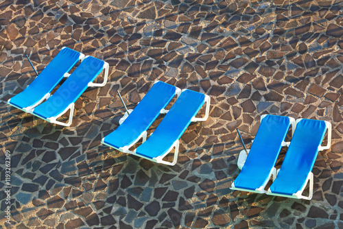 Blue lounge chairs arranged in a row on a patterned stone surface, likely by a poolside or a beach area. Chairs are positioned in such a way that they cast shadows on the ground
