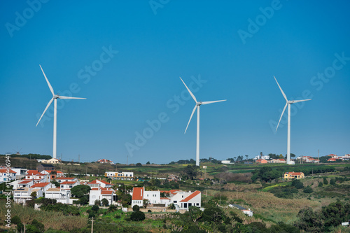 Wind turbines towering over a scenic countryside with white houses and green fields under a clear blue sky.
