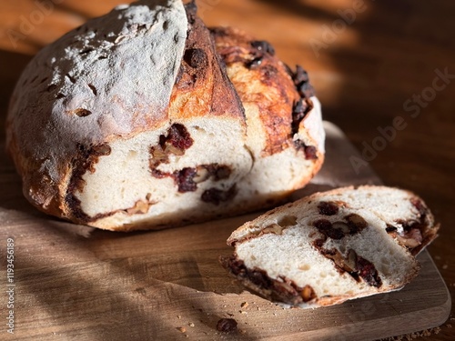 A sliced loaf of cranberry, walnut, molasses swirl sourdough bread. The loaf sits on a wooden cutting board, on a stained wood table, in warm sunlight. ..