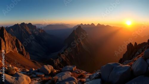 Sunset over Majestic Mountain Peaks, Golden Light Illuminating Rugged Landscape with Deep Blue to Soft Orange Sky Transition, Foreground Boulders, and Sparse Vegetation
