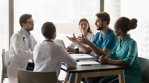 Young male doctor wear blue scrubs make speech during briefing with diverse clinic workmates, share knowledge, keep medical staff updated on latest research, technologies, treatment methods. Teamwork photo